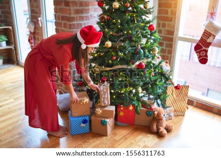 Young beautiful woman standing with happy face picking up present from under the christmas tree wearing santa claus hat