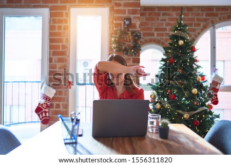 Beautiful woman sitting at the table working with laptop at home around christmas tree covering eyes with arm, looking serious and sad. Sightless, hiding and rejection concept