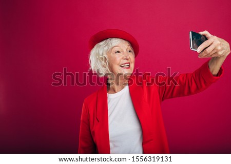 Attractive, positive and cheerful old woman take selfie using phone camera. Look at it and smile. Stylish senior model wear red coat and beret. Stand alone. Isolated over red background