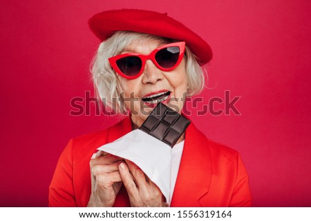 Portrait of old woman look straight through sunglasses and want to bite piece of chocolate. Wear stylish red beret and coat or sweater. Stand alone. Isolated over red background