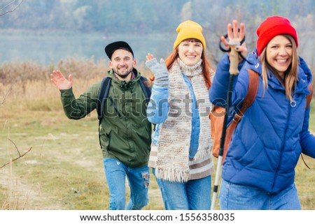 Smiling people travelers with backpacks and tracking poles waving, laughing walking in line in bright colorful clothes with monochrome late autumn naure on background. Positive emotions concept.