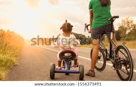 A picture of the warmth of a mother who is teaching her child to ride a bicycle, which the mother will take care to not be dangerous, public road in the evening with the orange light of the sunset.