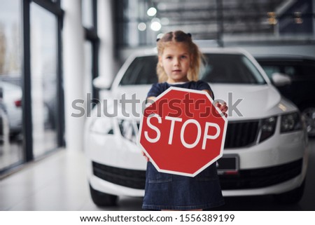 Portrait of cute little girl that holds road sign in hands in automobile salon.