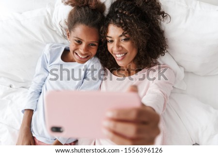 Smiling young mother and her little daughter wearing pajamas releaxing on bed, taking a selfie Royalty-Free Stock Photo #1556396126