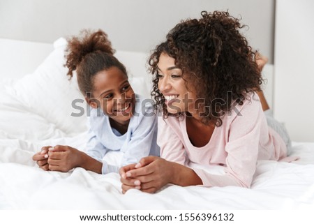 Smiling young mother and her little daughter wearing pajamas releaxing on bed, embracing Royalty-Free Stock Photo #1556396132