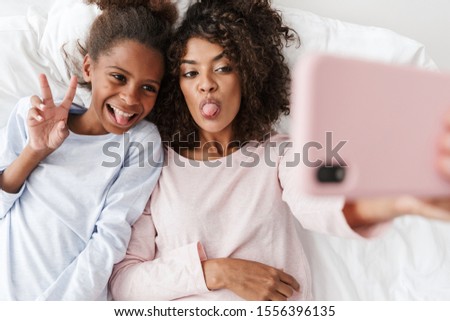 Smiling young mother and her little daughter wearing pajamas releaxing on bed, taking a selfie Royalty-Free Stock Photo #1556396135