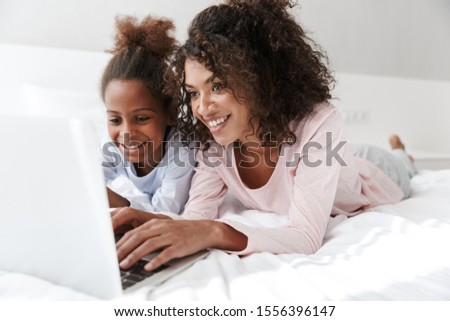 Smiling young mother and her little daughter wearing pajamas releaxing on bed, using laptop computer, chatting Royalty-Free Stock Photo #1556396147