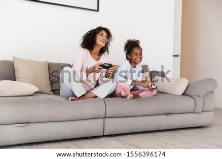 Smiling young mother and her little daughter wearing pajamas releaxing on a couch, playing video games Royalty-Free Stock Photo #1556396174