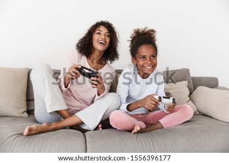 Smiling young mother and her little daughter wearing pajamas releaxing on a couch, playing video games Royalty-Free Stock Photo #1556396177