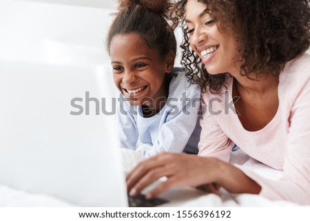 Smiling young mother and her little daughter wearing pajamas releaxing on bed, using laptop computer, chatting Royalty-Free Stock Photo #1556396192