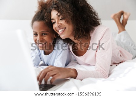 Smiling young mother and her little daughter wearing pajamas releaxing on bed, using laptop computer, chatting Royalty-Free Stock Photo #1556396195