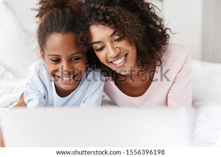 Smiling young mother and her little daughter wearing pajamas releaxing on bed, using laptop computer, chatting Royalty-Free Stock Photo #1556396198