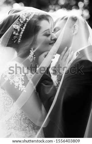Black and white photo. Wedding day. The pretty young bride and his groom are kissing under the bride's fate. Young wedding couple enjoying romantic moments.