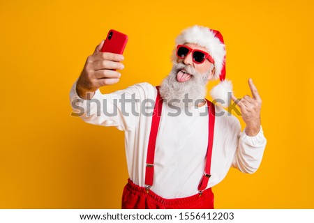 X-mas party hard! Portrait of funny crazy santa claus in red hat take selfie smart phone christmas blogger show horns grimace tongue out wear shirt suspenders isolated yellow color background