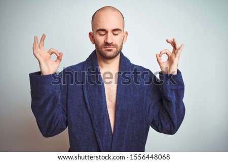 Young man wearing blue bathrobe, relaxed lifestyle over isolated background relax and smiling with eyes closed doing meditation gesture with fingers. Yoga concept.