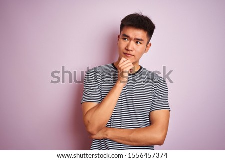 Young asian chinese man wearing striped t-shirt standing over isolated pink background with hand on chin thinking about question, pensive expression. Smiling with thoughtful face. Doubt concept.