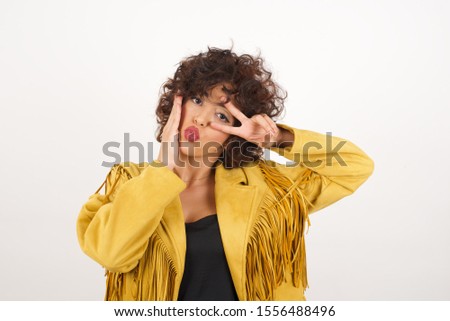 Leisure lifestyle people person celebrate flirt coquettish concept. Beautiful brunette woman wearing fringed jacket showing making v-sign near eyes wearing casual clothes standing against white wall.