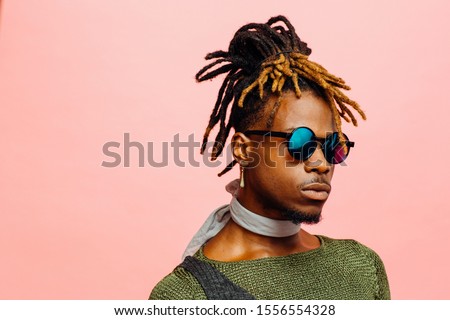 Close up portrait of a young man in green with dreadlocks and blue sunglasses, isolated on pink Royalty-Free Stock Photo #1556554328