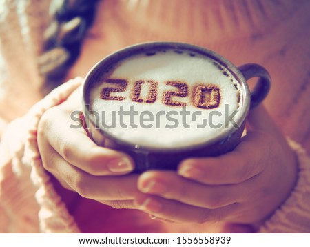 Number 2020 on frothy surface of cappuccino served in white cup holding by female hands with French nail polish. Food art creative concept for active days in New Year 2020. (close up, selective focus) Royalty-Free Stock Photo #1556558939