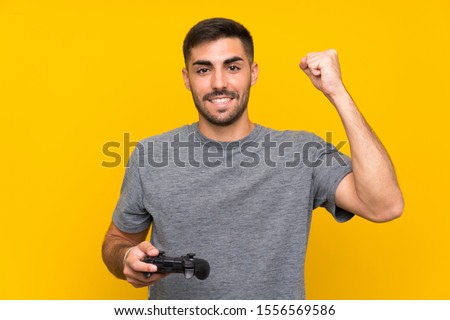 Young handsome man playing with a video game controller over isolated yellow background celebrating a victory