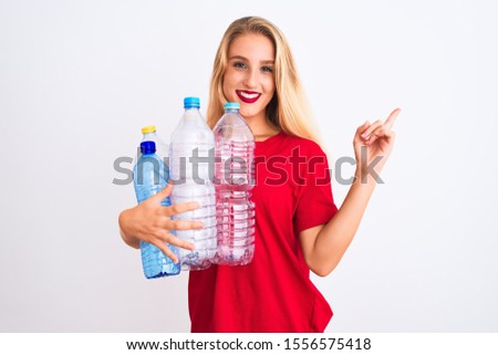 Young beautiful woman recycling plastic bottles standing over isolated white background very happy pointing with hand and finger to the side