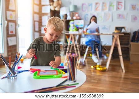 Young caucasian child playing at playschool with teacher. Mother and son at playroom drawing a draw with color pencils, young woman at the background sitting on desk.