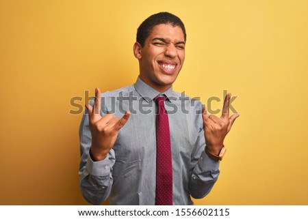 Young handsome arab businessman wearing shirt and tie over isolated yellow background shouting with crazy expression doing rock symbol with hands up. Music star. Heavy concept.