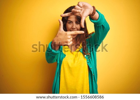 Young woman wearing t-shirt and green sweater standing over yelllow isolated background smiling making frame with hands and fingers with happy face. Creativity and photography concept.