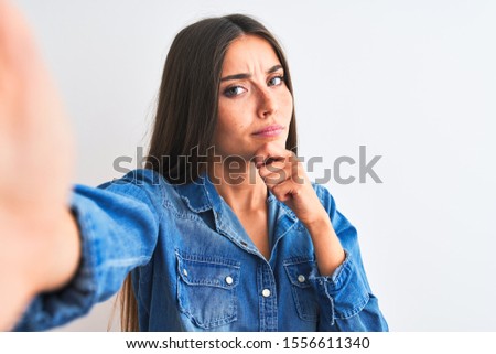 Beautiful woman wearing denim shirt make selfie by camera over isolated white background looking confident at the camera smiling with crossed arms and hand raised on chin. Thinking positive.