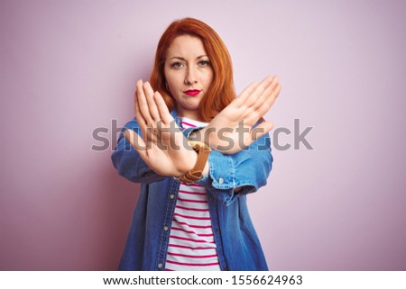 Beautiful redhead woman wearing denim shirt and striped t-shirt over isolated pink background Rejection expression crossing arms and palms doing negative sign, angry face