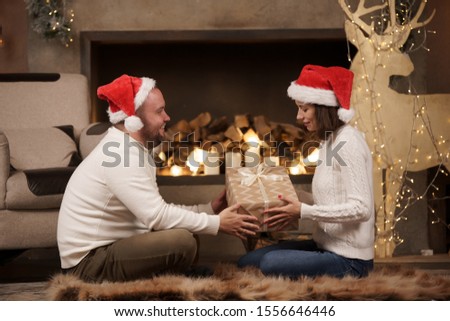 Photo from side of man and woman in Santa's caps sitting on floor in room