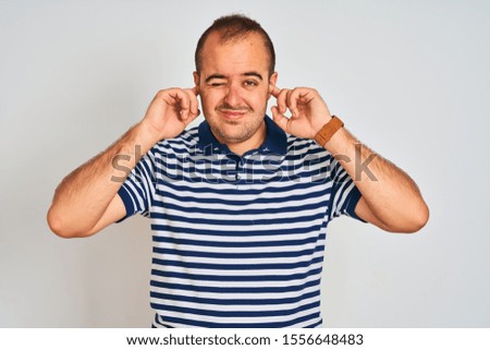 Young man wearing casual striped standing over isolated white background covering ears with fingers with annoyed expression for the noise of loud music. Deaf concept.