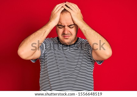 Young man wearing striped navy t-shirt standing over isolated red background suffering from headache desperate and stressed because pain and migraine. Hands on head.