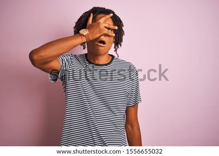 Afro man with dreadlocks wearing navy striped t-shirt standing over isolated pink background peeking in shock covering face and eyes with hand, looking through fingers with embarrassed expression.