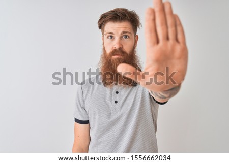 Young redhead irish man wearing grey shirt standing over isolated white background doing stop sing with palm of the hand. Warning expression with negative and serious gesture on the face.
