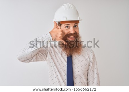 Young redhead irish architect man wearing security helmet over isolated white background smiling doing phone gesture with hand and fingers like talking on the telephone. Communicating concepts.