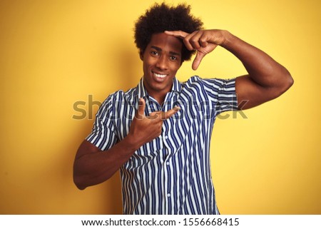 American man with afro hair wearing striped shirt standing over isolated yellow background smiling making frame with hands and fingers with happy face. Creativity and photography concept.