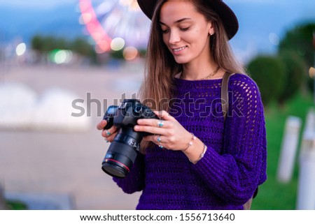 Happy beautiful casual stylish hipster traveler woman photographer in hat looking photos on dslr camera after taking pictures while walking around the city