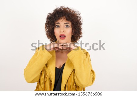 Pretty surprised lady in white pants fooling around in studio and jumping. Adorable girl in knitted sweater dancing on colorful background and touching chin with hands