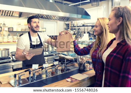 Waiter serving takeaway food to customers at counter in small family eatery restaurant – Smiling owner of trendy popular fast food delivering to clients a online to go order in recycled paper bag   Royalty-Free Stock Photo #1556748026