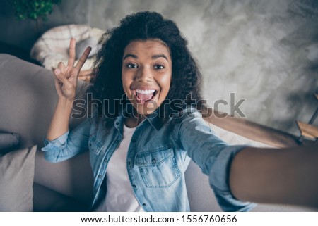 Closeup photo of pretty funky dark skin curly blogger lady making selfies excited showing v-sign symbol sitting comfy couch casual denim outfit living room indoors