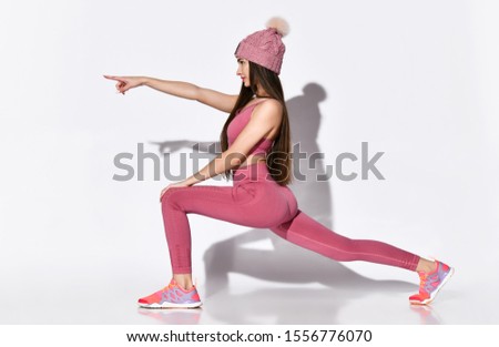 picture of an energetic brunette athlete of the 20s in a tracksuit in pink top and legends, a warm knitted hat with a pompom, lunges and stretching her legs, shows a hand in the direction of the look