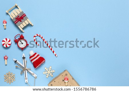 Corner creative Christmas composition. Candy cane, gift in craft paper, sled with deer, hat, alarm clock, ski, clothespins, wooden snowflakes on blue background, copy space. Minimal style. Top view.
