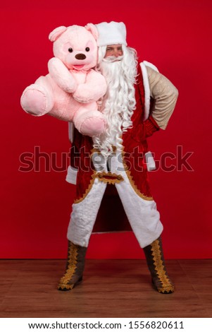 Emotional Santa Claus with a long white beard in a red coat and white hat posing with a big teddy pink bear on a red background