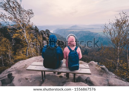 Young couple in outdoor clothing with backpacks sitting and rest on bench enjoying view of mountain ridge, forest and river in the valley on hiking trail. Travel lifestyle concept