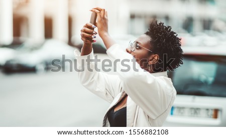 Side view of a young African female outdoors in a white cloak and spectacles holding a smartphone in her hands with nail-art and taking pics of something above using a rear camera of the gadget