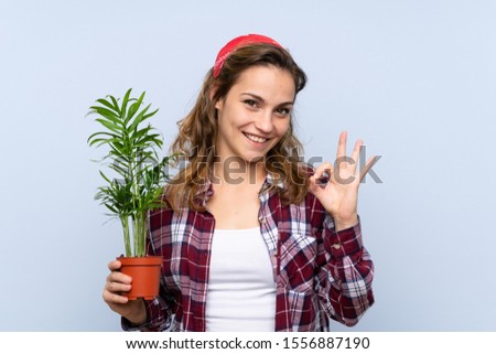 Young blonde gardener girl holding a plant showing ok sign with fingers