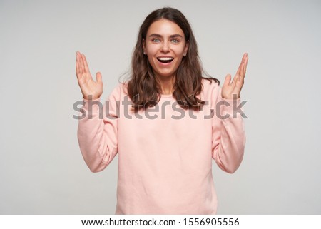 Portrait of cheerful pretty blue-eyed young female with dark hair looking happily to camera with wide sincere smile, posing over white background with raised hands