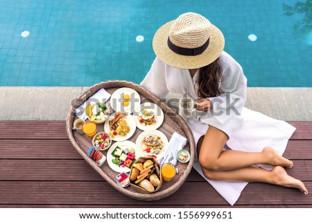 Served floating breakfast in 5 star hotel resort. Pretty young woman enjoying breakfast in the pool at luxury villa. Beautiful brunette girl relaxing and drinking coffee Royalty-Free Stock Photo #1556999651
