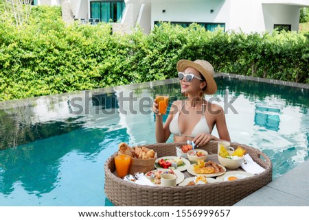 Served floating breakfast in 5 star hotel resort. Pretty young woman enjoying breakfast in the pool at luxury villa. Beautiful brunette girl relaxing and drinking coffee Royalty-Free Stock Photo #1556999657